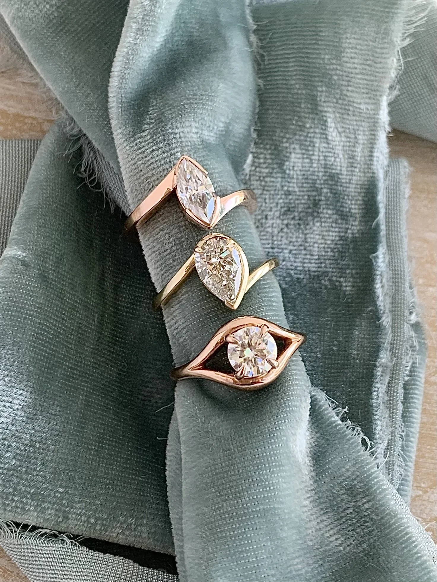 Three solitaire rings resting on sage velvet: a bezel-set marquise in rose gold, a bezel-set pear in yellow gold, and a round diamond in a rose gold split shank